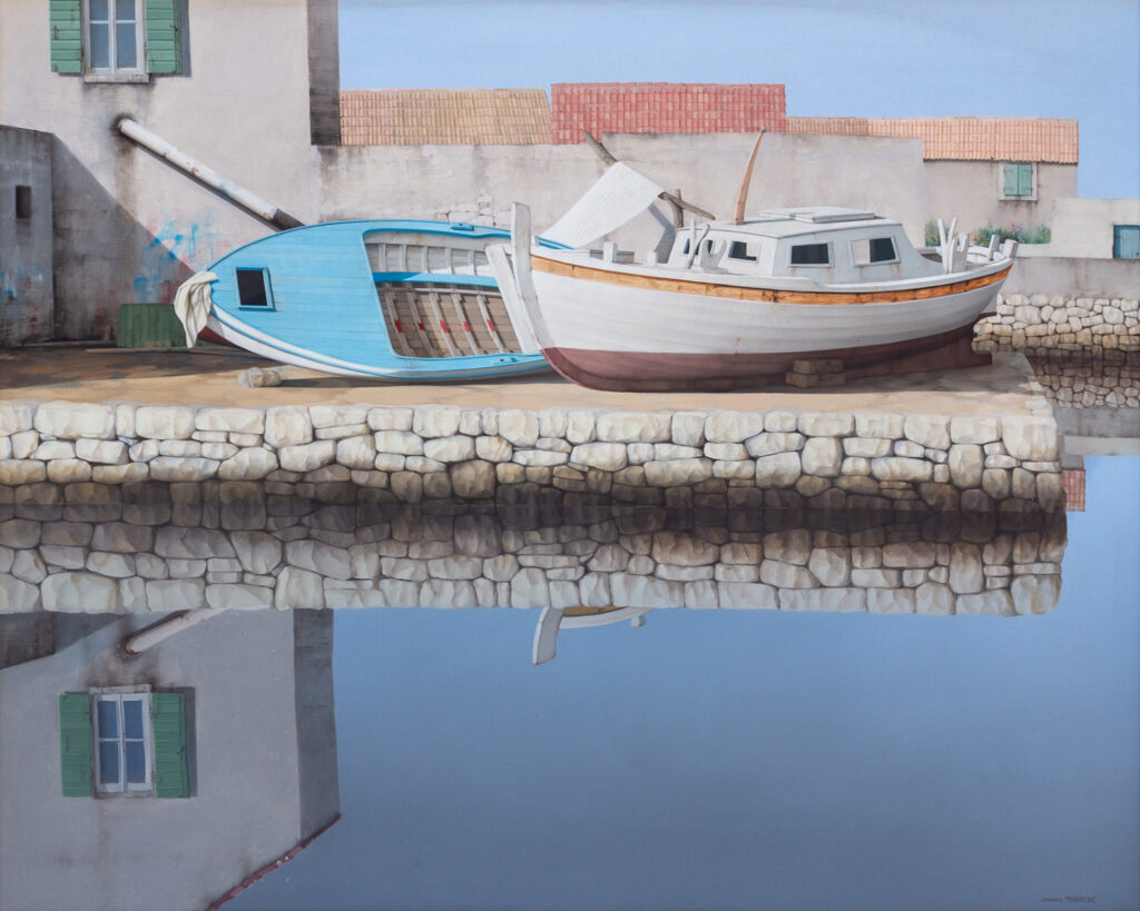Two wooden boats resting on a stone dock beside a weathered building with green shutters, reflecting both boats and building in calm, still water under a clear sky.